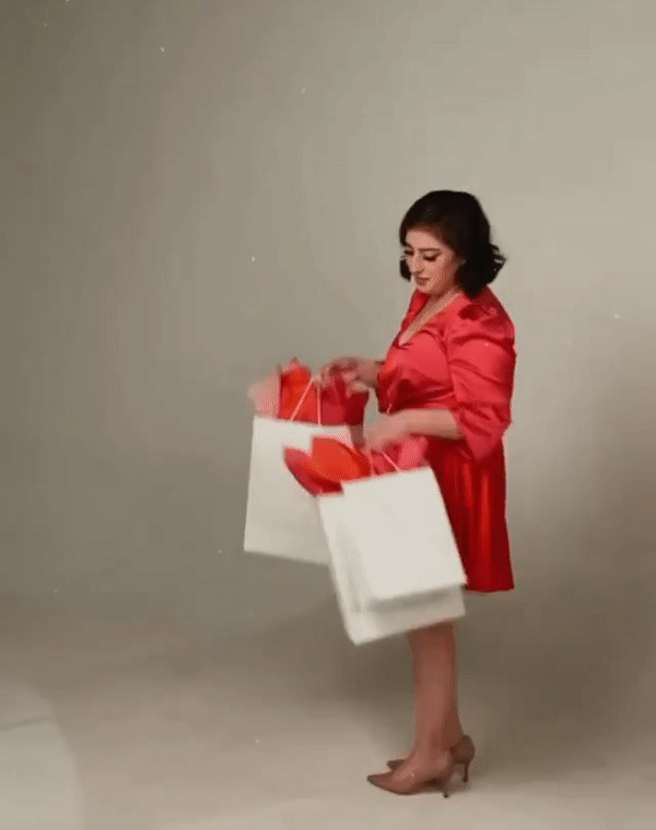 GIF of Yasamin Salavatian holding up paper shopping bags filled with red packaging paper. She is wearing a red dress.
